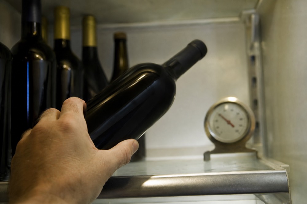 A hand placing a dark wine bottle onto a refrigerator shelf beside a kitchen thermometer