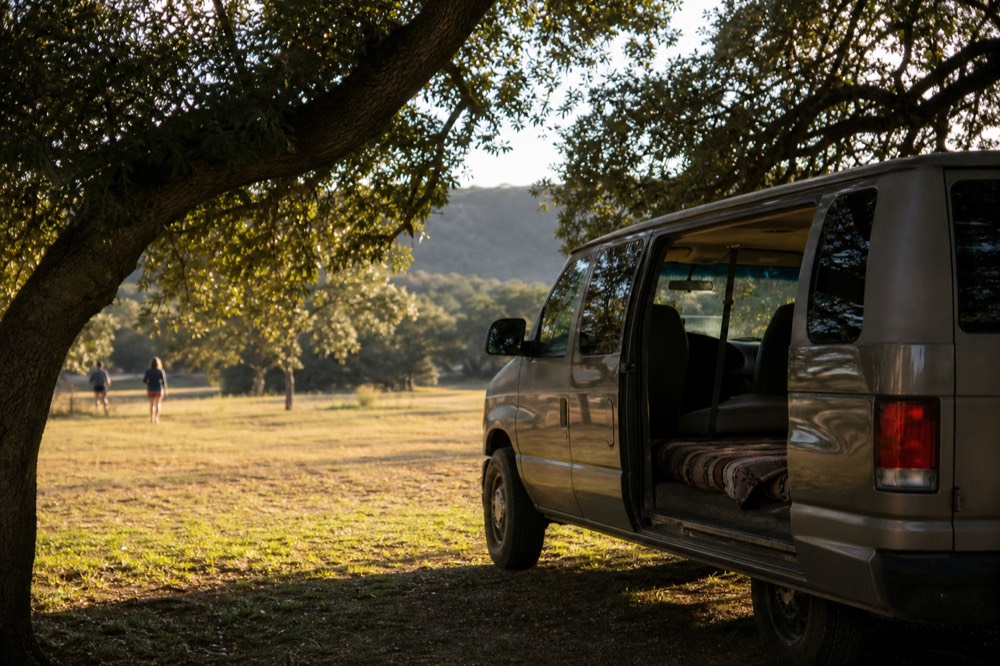 An enclosed passenger van parked in shade with its side door open and a folded blanket on the rear floor