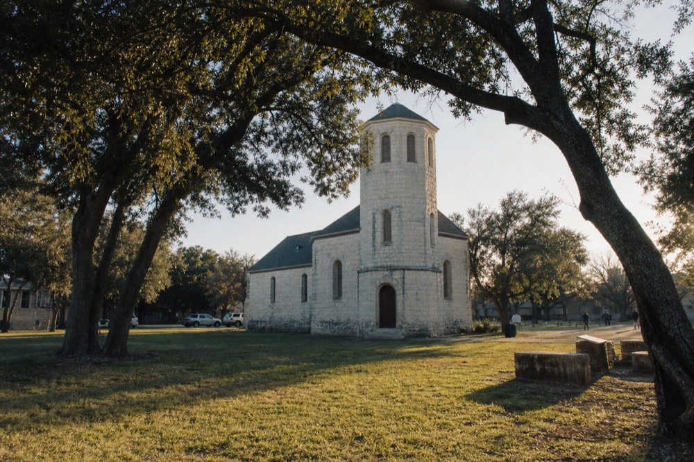 A white limestone church-like building with a hexagonal tower standing alone in the middle of an open town square, mature trees around the edges