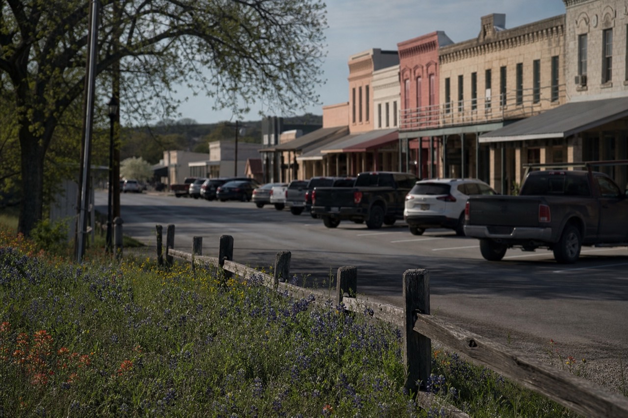 A wide small-town main street lined with low limestone and painted-brick storefronts under deep awnings, angled parking, warm afternoon light