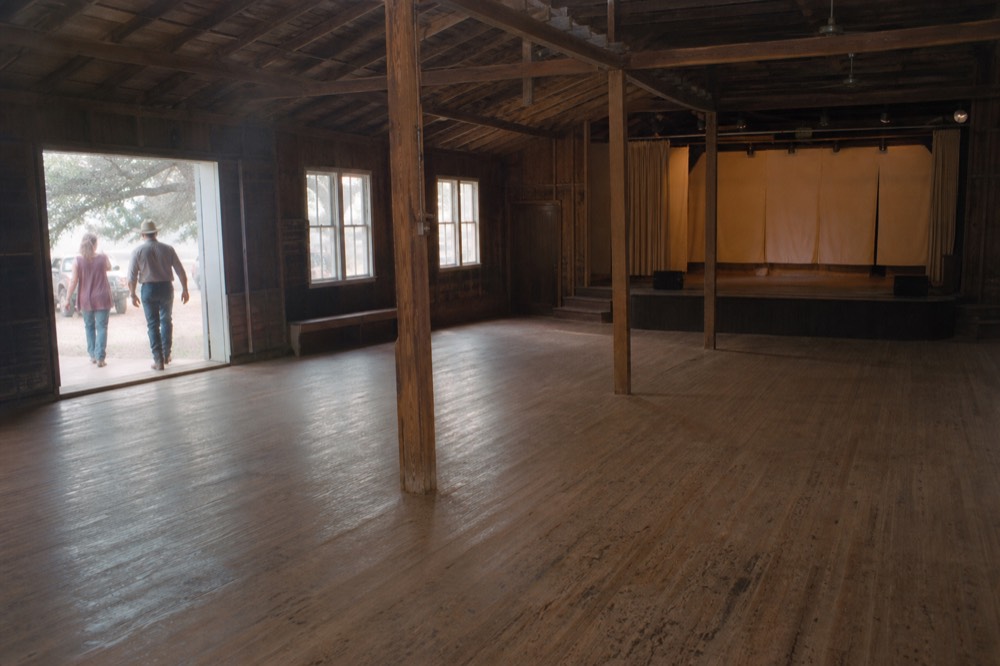 An old wooden dance hall interior with a worn plank floor, exposed rafters and a small empty stage at the far end