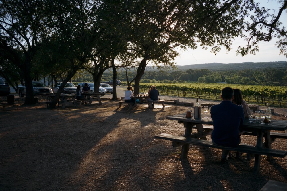 Long shadows across a dirt yard of picnic tables under oak trees, a few people sitting with drinks in late golden light