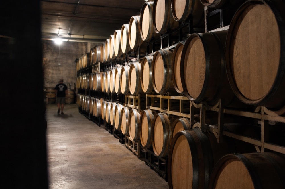 A long line of stacked oak barrels in a dim cellar with a concrete floor and a single work light overhead