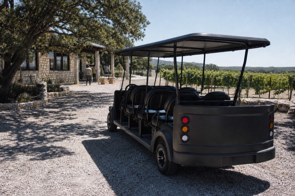 A small open-air touring vehicle parked outside a vineyard tasting room with rows of vines behind and gravel underfoot