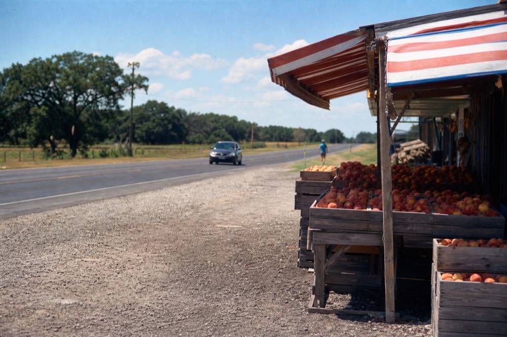 Roadside peach stand with crates of fruit under a striped awning beside a rural Texas highway in summer