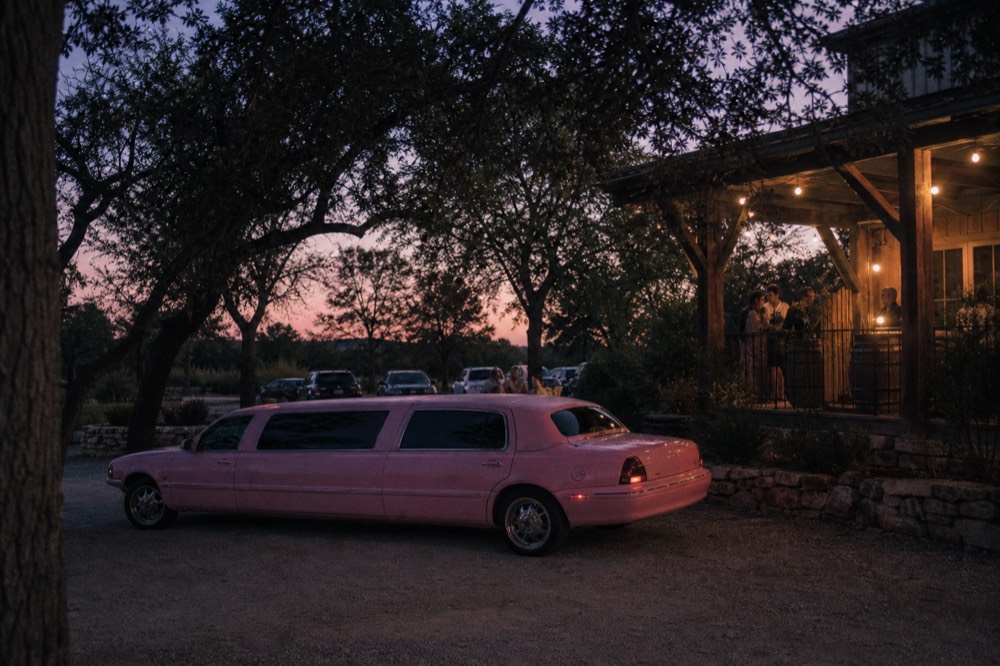 A stretched pastel-pink compact limousine parked on a gravel drive under live oaks, no branding or plates visible
