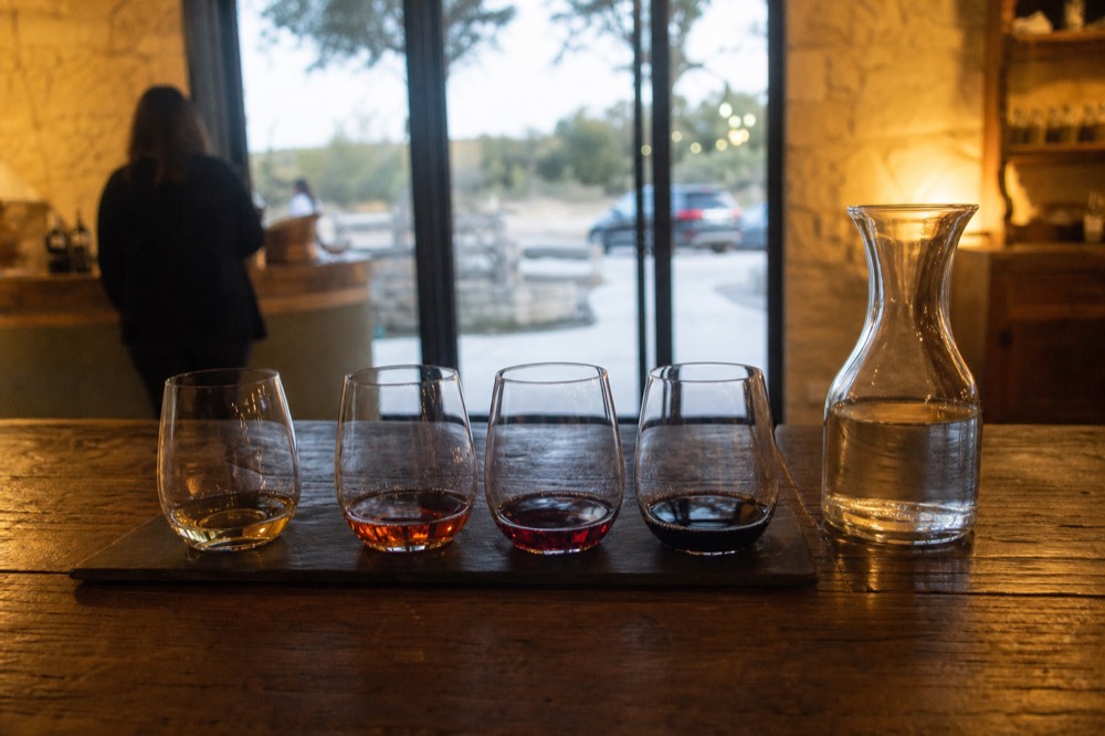 A wooden tasting bar with four small pour glasses lined up on a slate flight board, a pitcher of water beside them, warm interior light