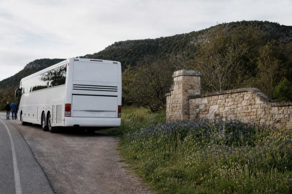 A small touring coach on a rural highway shoulder beside a stone winery entrance wall, hills rising behind