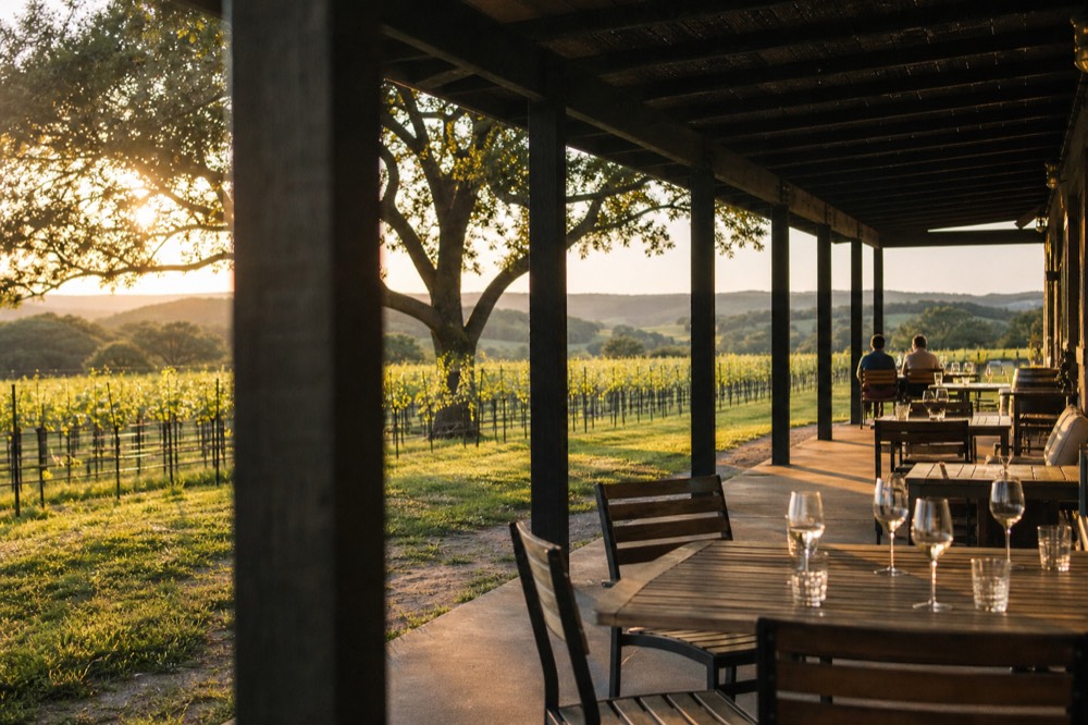 A long shaded tasting-room patio with wooden tables and scattered wine glasses looking out over vine rows and low hills