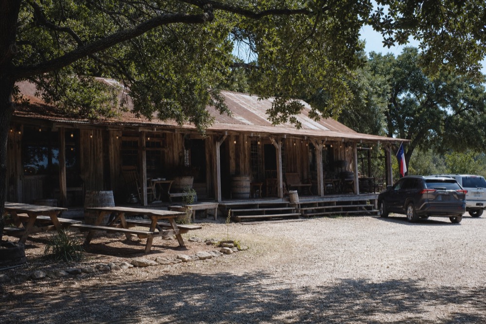 A weathered wooden general store with a covered porch under large live oaks, string lights overhead and picnic tables in the dirt yard