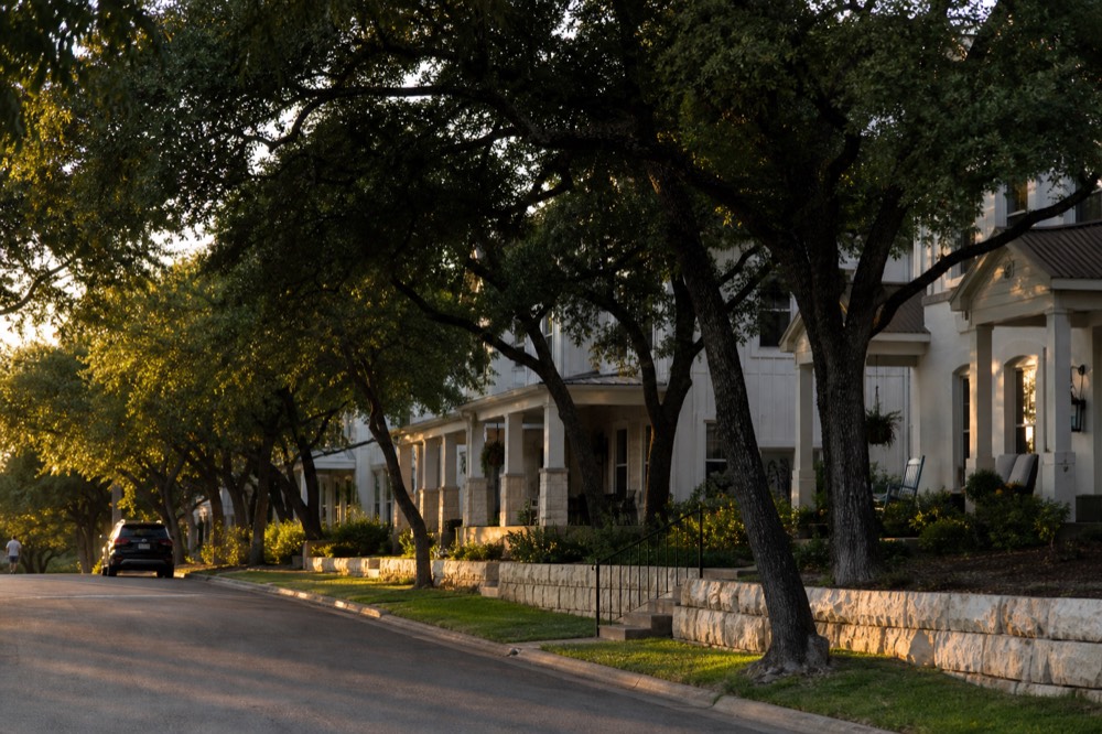 A quiet residential street of white limestone cottages with deep porches under mature oaks, early evening