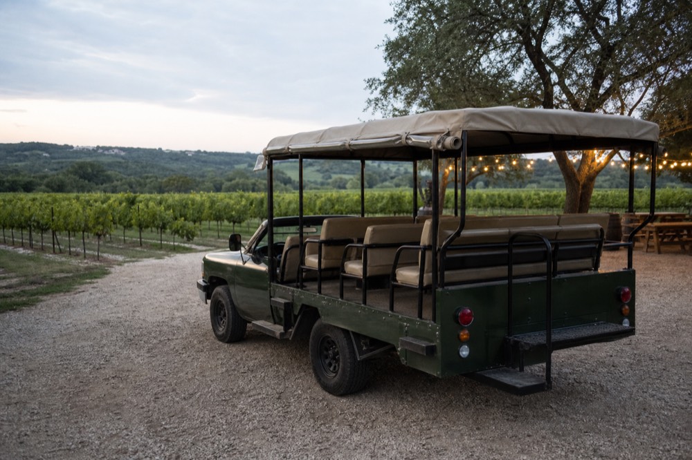 An open-air tour vehicle parked on gravel beside rows of grapevines, empty bench seats and a canvas roof, mid-morning