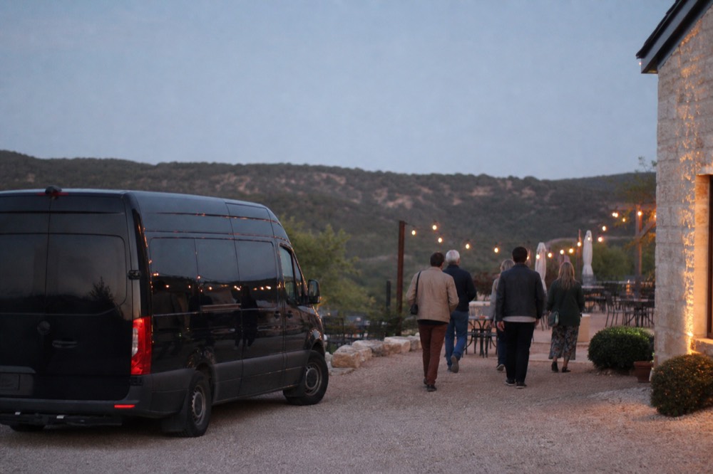 A black Sprinter van parked at the front entrance of a winery while a small group walks toward a reserved outdoor table set with glasses