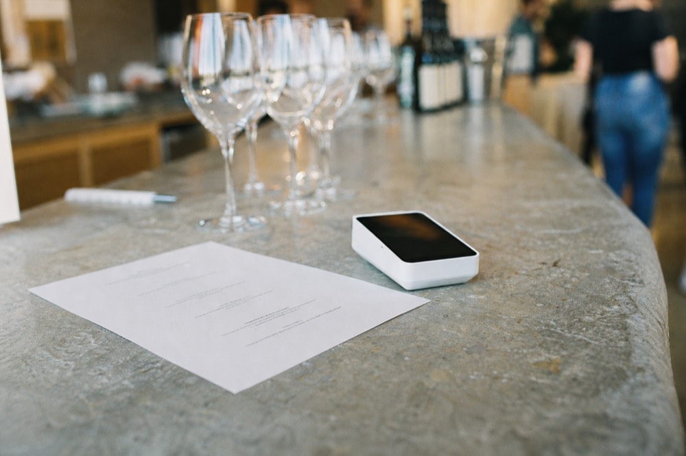 Close-up of a printed tasting menu and a card reader on a limestone winery bar, glasses out of focus behind
