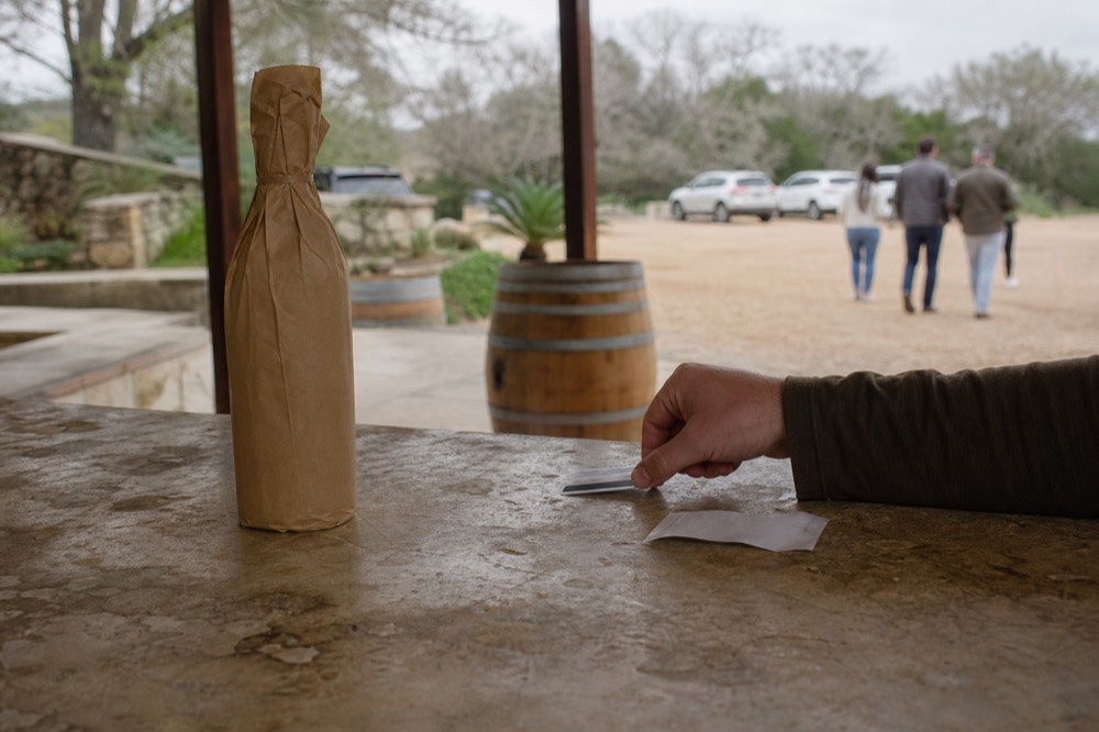 Hand sliding a credit card across a winery counter beside a wrapped bottle and a receipt