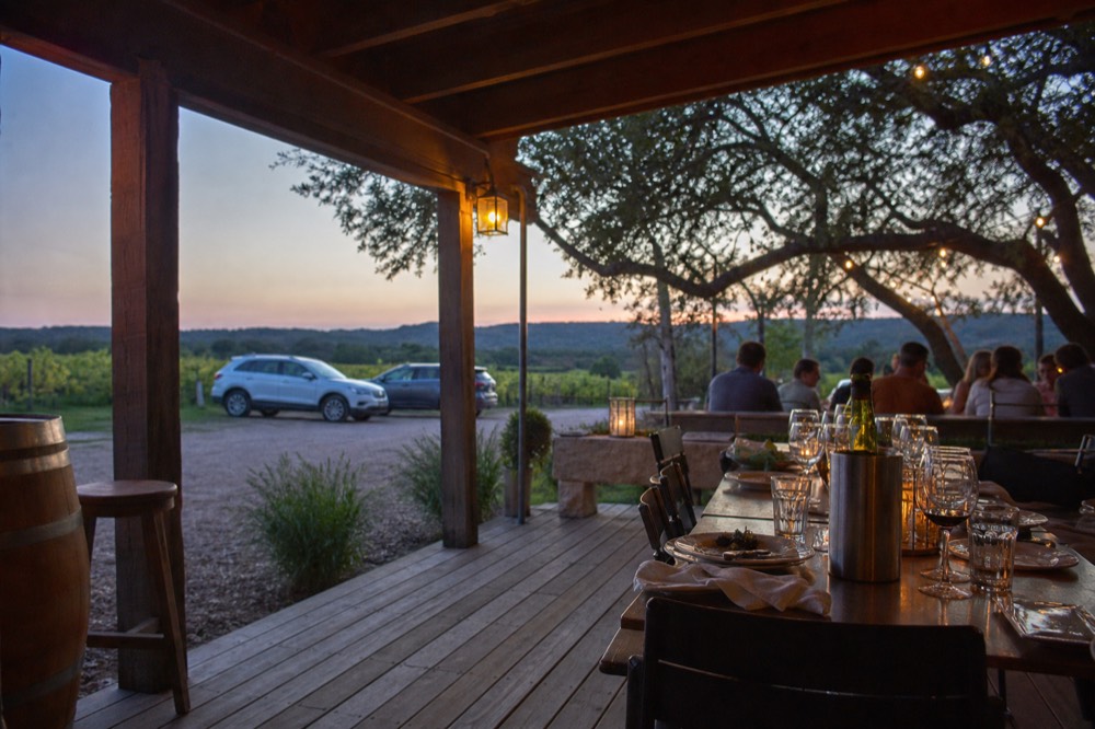 Outdoor lunch table set with plates and wine glasses under an oak tree at a Texas vineyard, guests seated in the background