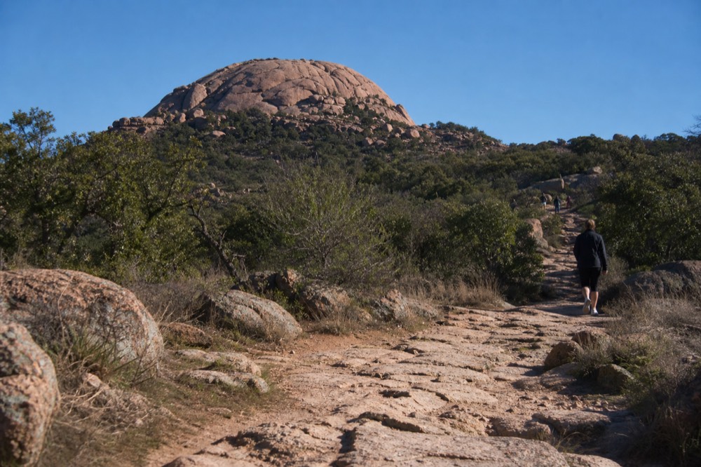 A pink granite dome rising above scrub oak at sunrise with a rough hiking trail in the foreground